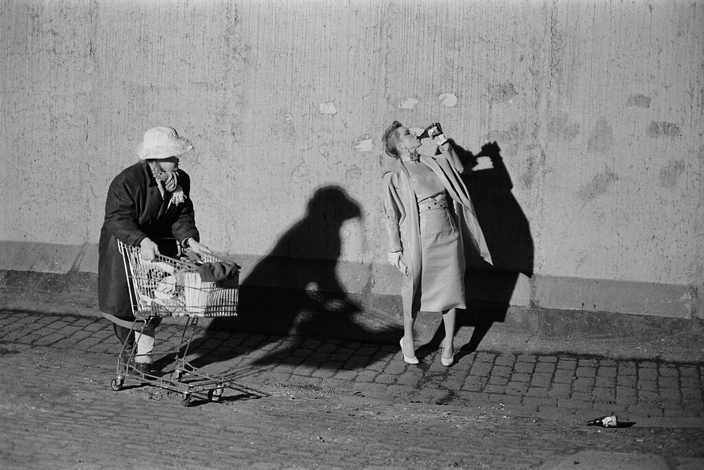 Lady with a shopping cart watching another lady drinking from a bottle on a street.