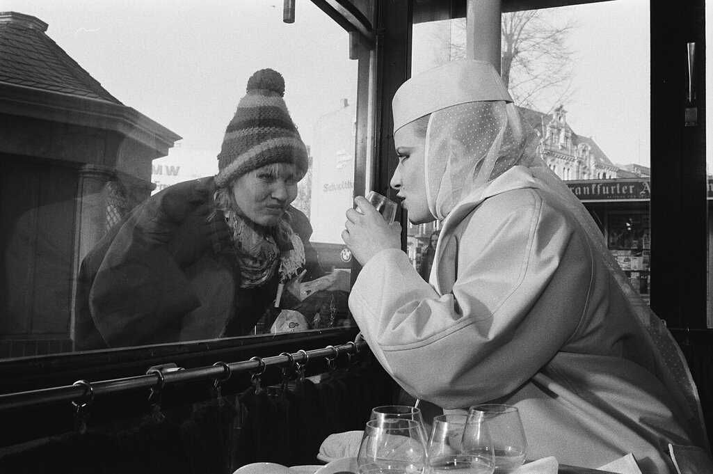 A woman gazing through a window at another lady drinking a drink.